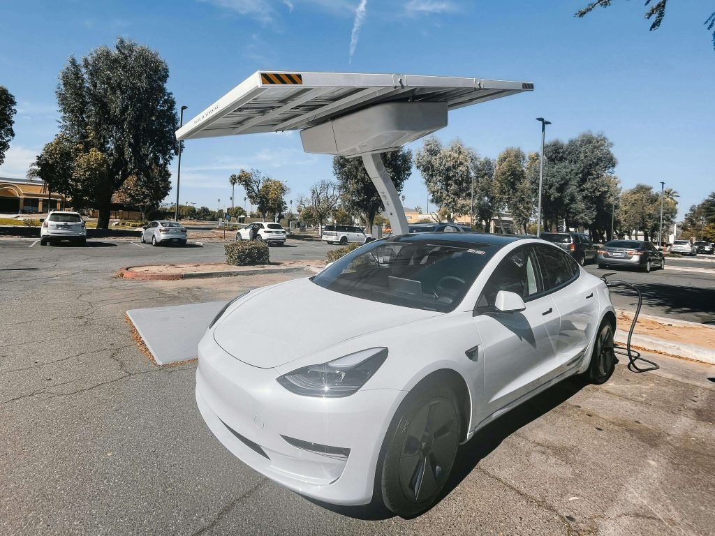 A white electric car charges under a solar panel canopy in a parking lot. Surrounded by parked cars and trees, the scene feels modern and eco-friendly.
