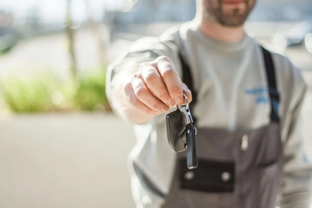 Close-up of a person handing car keys forward, wearing a light-colored shirt and dark overalls. The background is blurred, creating a professional tone.