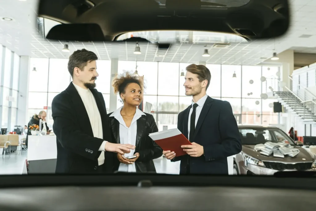 Three people in formal attire stand in a bright car showroom, engaged in conversation. The setting is modern and open, with cars visible behind them.