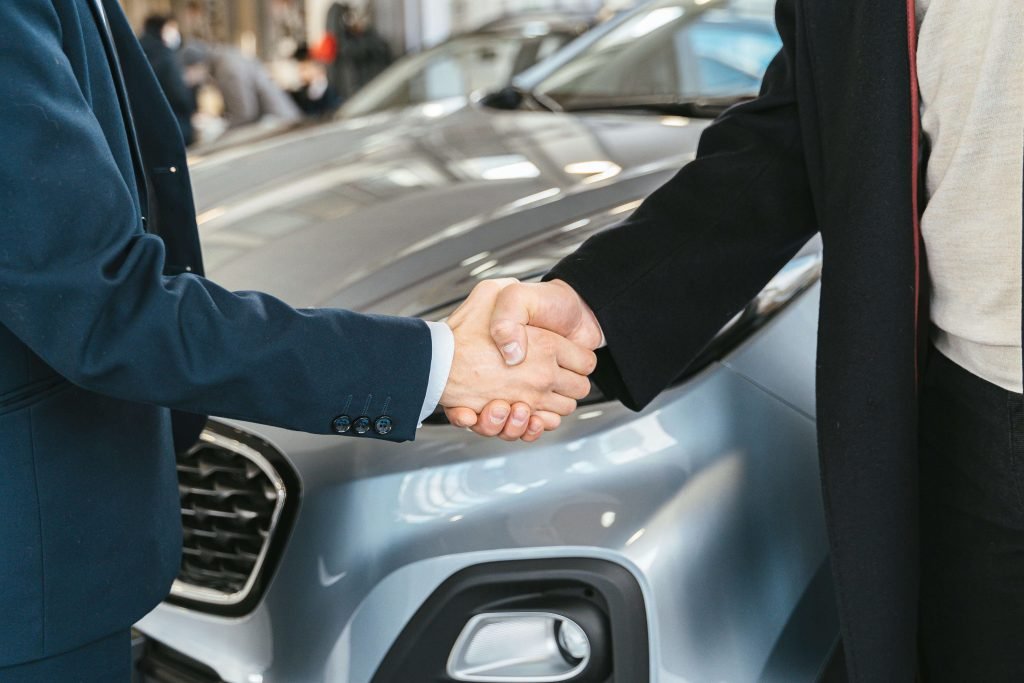 Two people in business attire are shaking hands in front of a silver car, indicating a successful transaction or agreement in a car dealership setting.