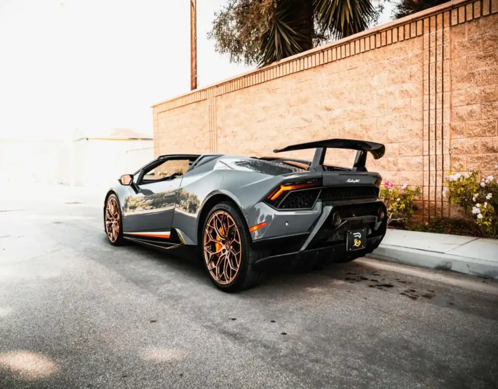 A sleek gray sports car with bronze rims and a bold rear spoiler is parked by a sunlit brick wall, exuding luxury and performance vibes.
