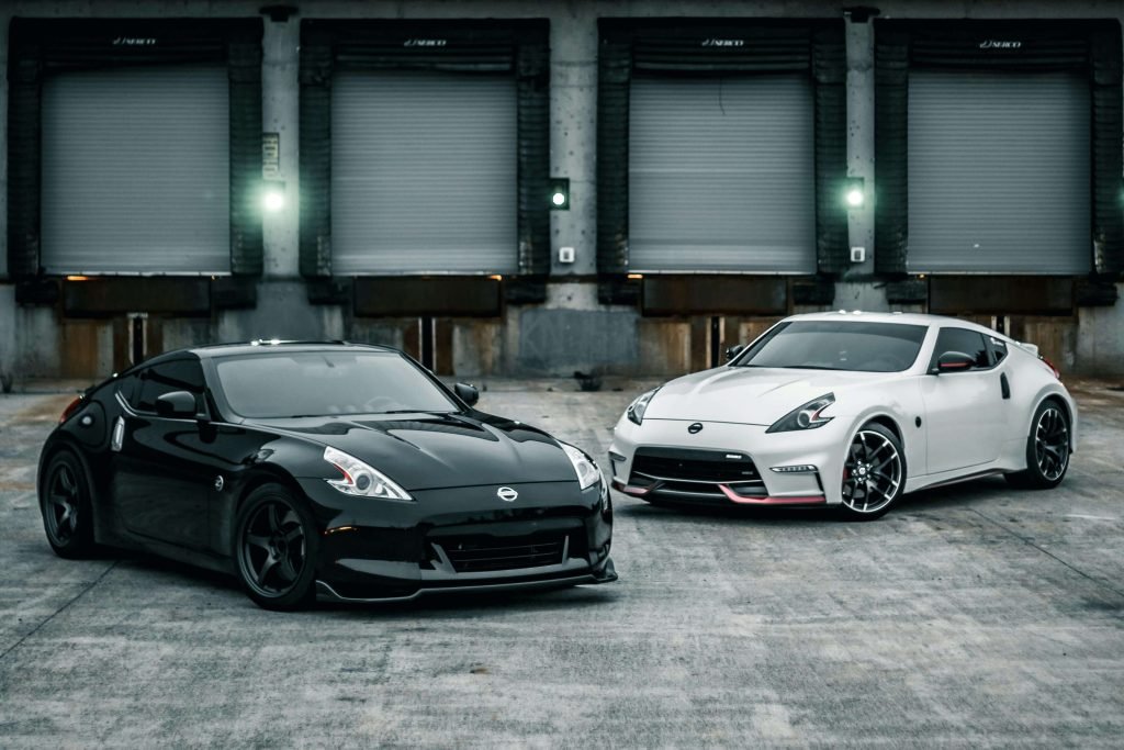 Two sleek sports cars, one black and one white, positioned on a concrete surface in front of industrial loading docks. The scene feels modern and dynamic.