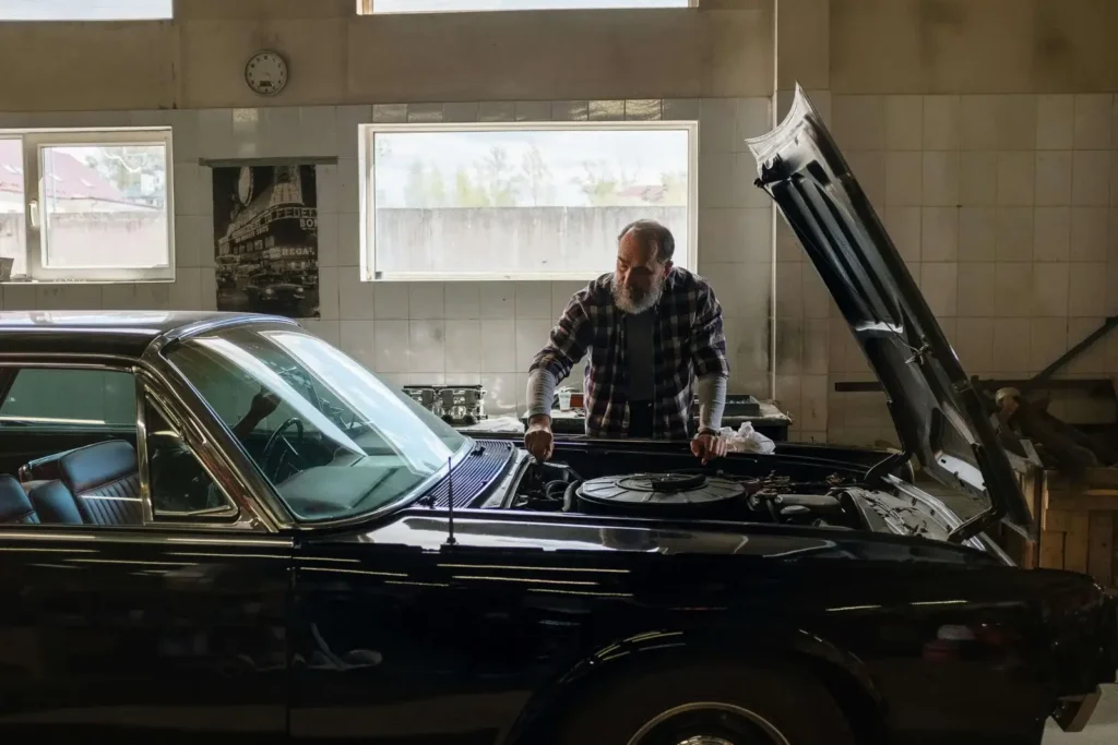 An older man with a beard is examining the engine of a classic black car with the hood open inside a workshop. Natural light enters through large windows.