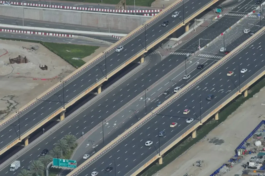 Aerial view of a multi-lane highway with moderate traffic, featuring multiple bridges and overpasses, surrounded by sandy and grassy areas.