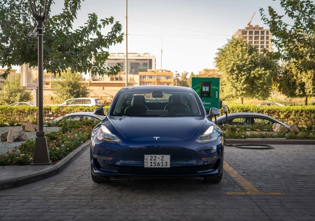 Blue Tesla Model 3 charging at a green station, framed by trees and city buildings in the background. Bright, clean, and modern urban setting.