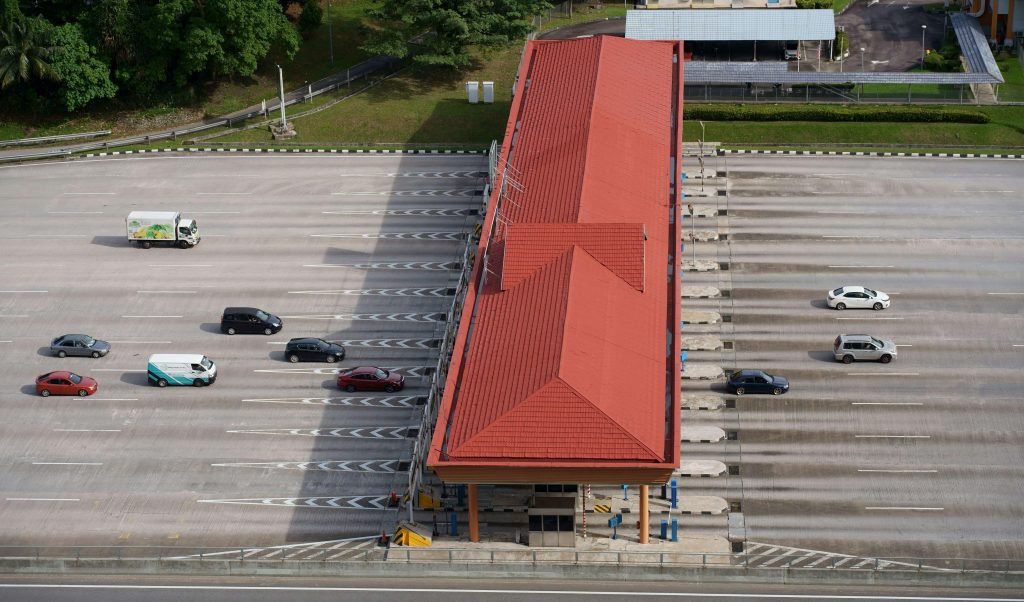 Aerial view of an empty red-roofed toll booth with several cars bypassing it on a wide road, surrounded by greenery. The mood is calm and orderly.