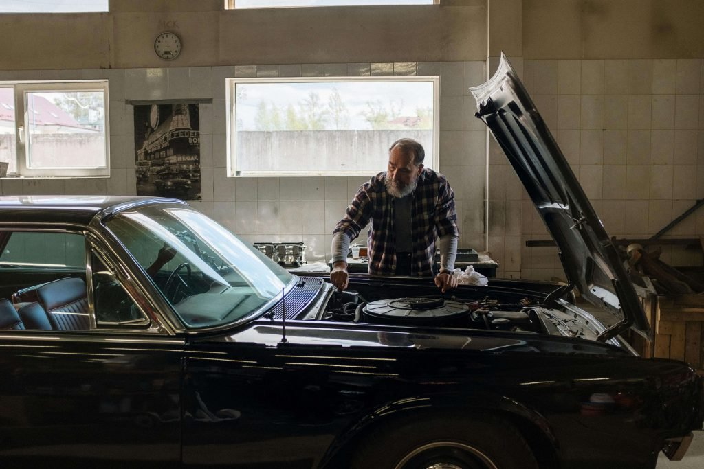 An older man with a beard is examining the engine of a classic black car with the hood open inside a workshop. Natural light enters through large windows.