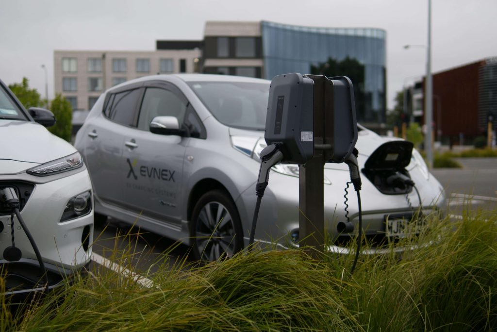 Two electric cars are parked and charging at a station in an urban setting. Green grass is in the foreground, and modern buildings are visible behind.