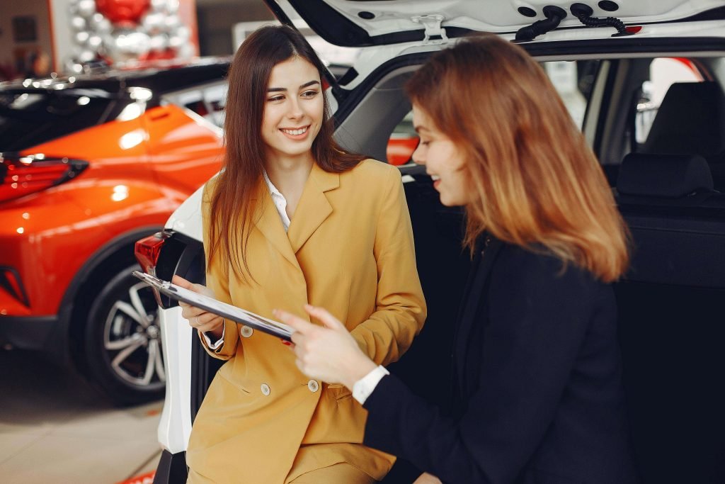 Two women smiling and conversing at the open trunk of a car in a showroom. One holds a clipboard. Background features red and black cars.