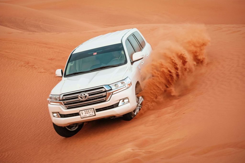 A white SUV drives through desert sand dunes, creating a spray of red sand. The dynamic motion conveys adventure and excitement.