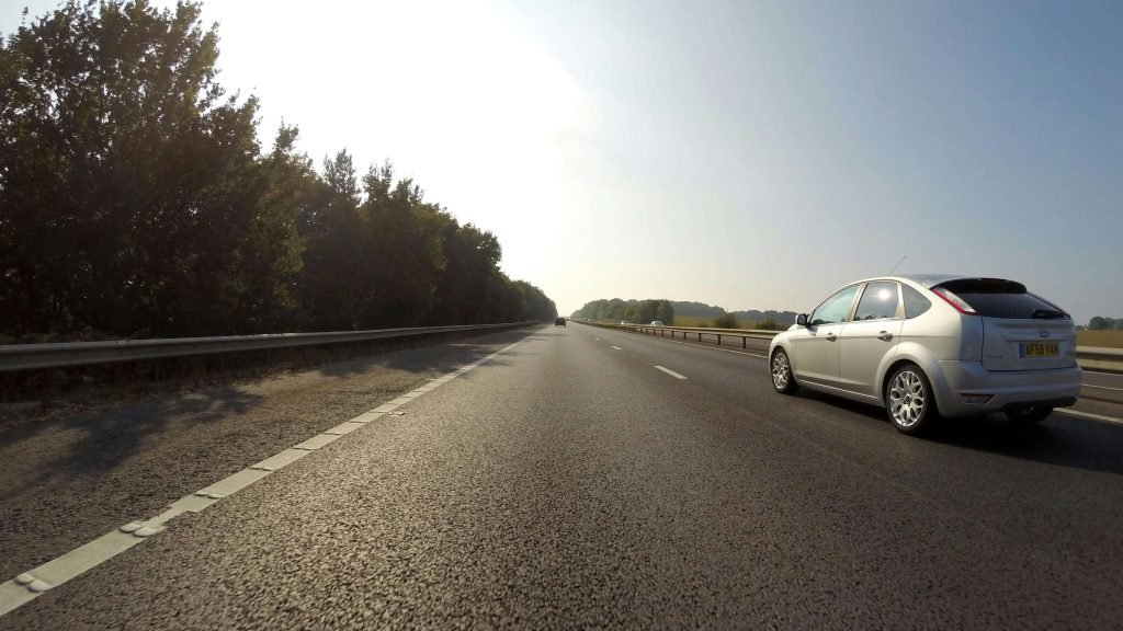 A silver car drives on an empty highway bordered by trees under a bright sky. The scene conveys a sense of freedom and open road adventure.
