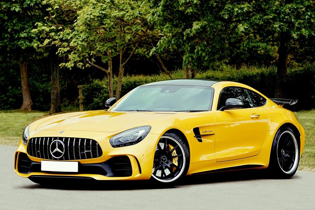 A sleek yellow sports car, parked on a road, framed by lush green trees. The car has a low profile and shiny alloy wheels, conveying speed and luxury.