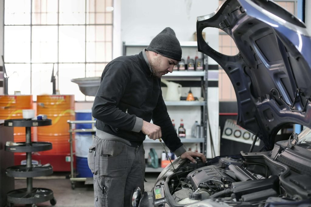 A mechanic in a dark jacket and beanie inspects a car engine in a garage. The hood is open, revealing engine parts. Tools and products are in the background.