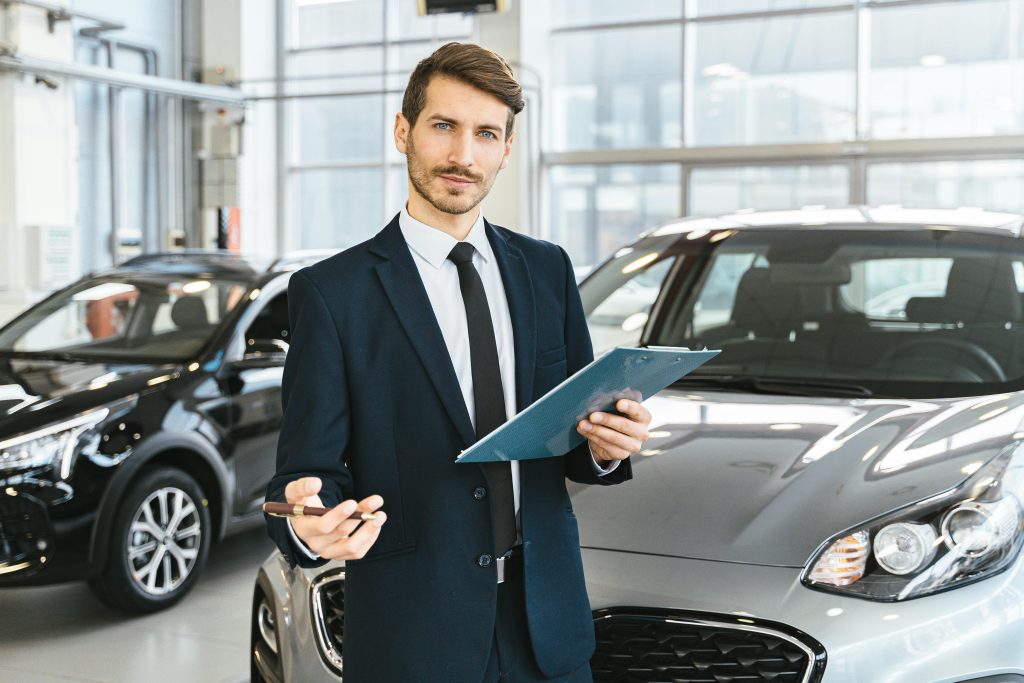 A man in a suit stands in a car showroom, holding a clipboard and pen, looking confident. Two modern cars are parked behind him in a bright, spacious area.