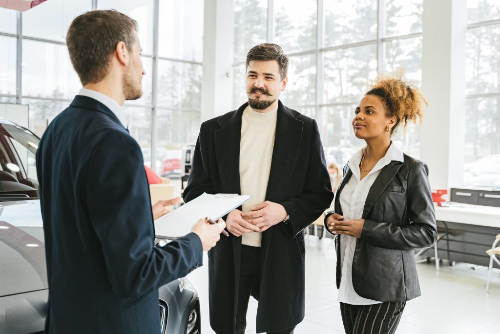 A couple consults with a salesperson in a bright car showroom. The man holds a clipboard while the couple listens attentively, expressing interest.