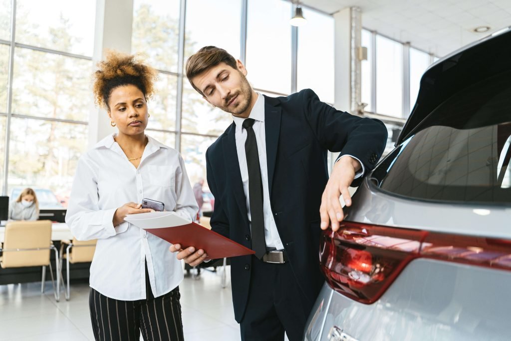 A man in a suit shows a woman the rear of a car in a bright showroom. She holds a phone and paperwork, appearing attentive and engaged.