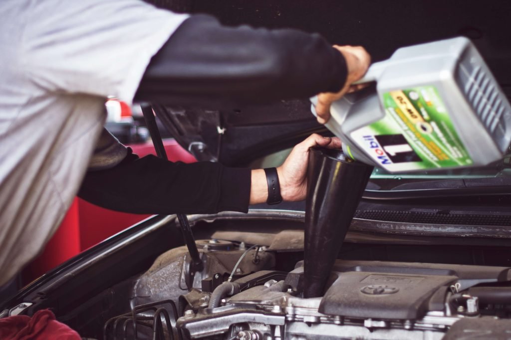 A mechanic wearing a black and white shirt pours motor oil into a car engine using a funnel. The focus is on the action of oil maintenance.