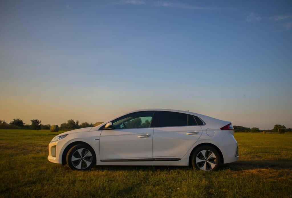 A white hatchback car is parked on a grassy field under a clear blue sky during sunset. The warm, golden light creates a serene atmosphere.