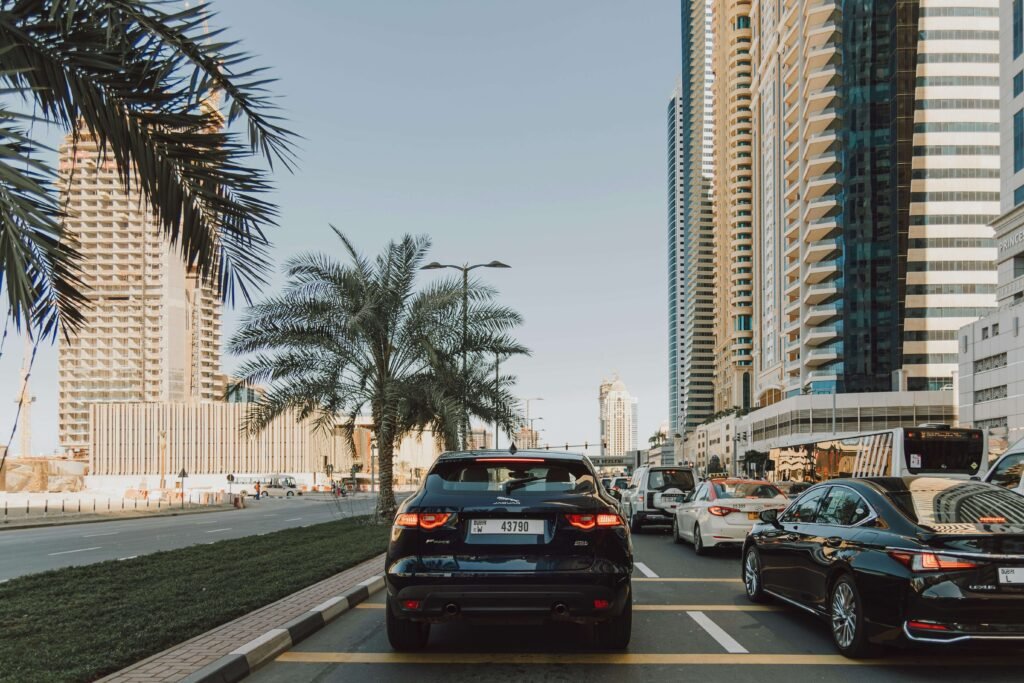 A busy city street under a clear sky, with cars queued in traffic. Tall skyscrapers line the road, and palm trees add a touch of greenery.