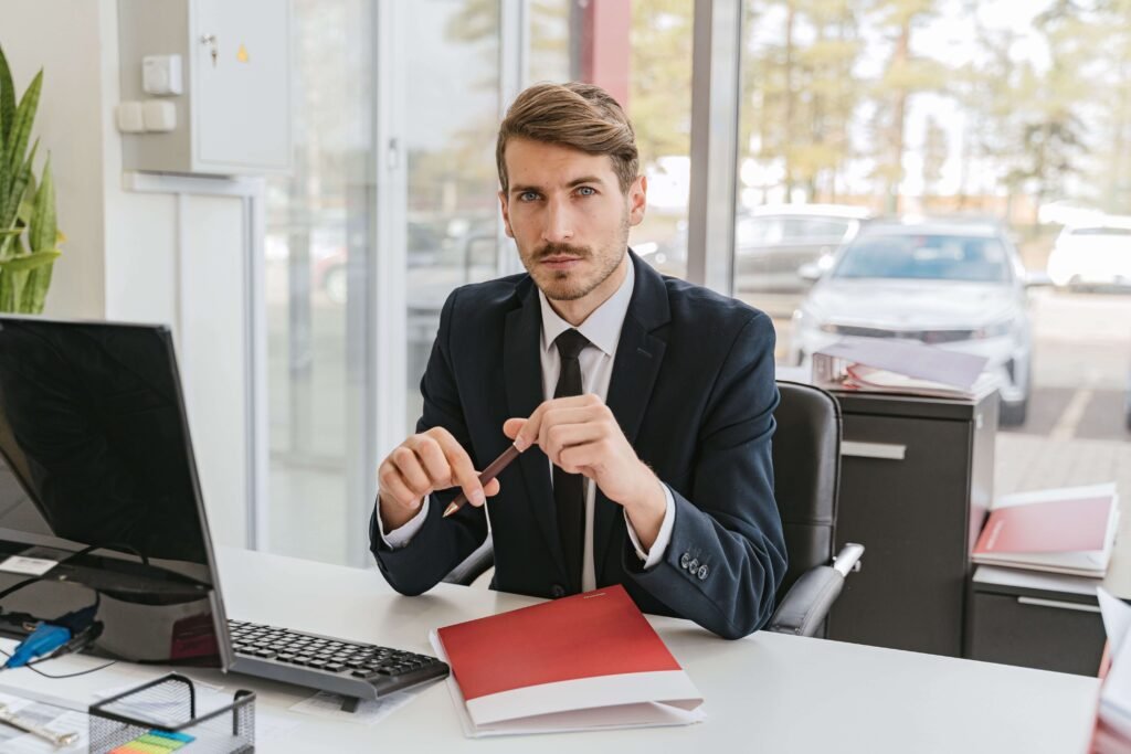 A man in a suit sits at a desk with a laptop and folder in a bright office, looking serious. Cars and trees are visible through the window behind him.