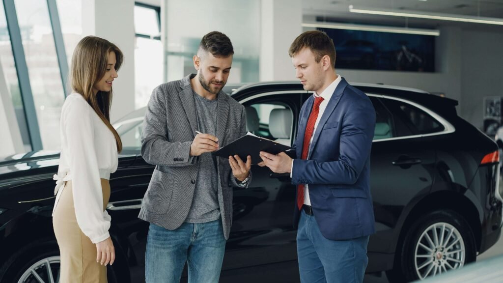 A man in a blazer and jeans signs a document on a clipboard held by a man in a suit and red tie, while a woman watches, beside a black car in a showroom.
