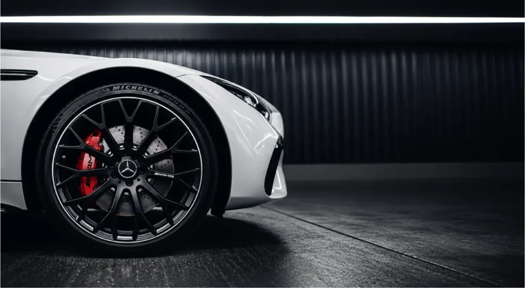 Close-up of a sleek white luxury sports car, featuring a black alloy wheel, red brake caliper, and Mercedes emblem in a dark garage.